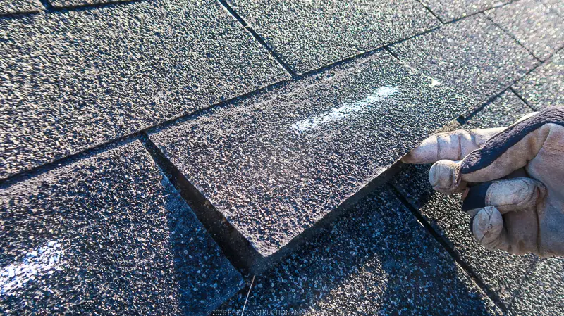 A gloved hand lifting an asphalt shingle during a roof inspection