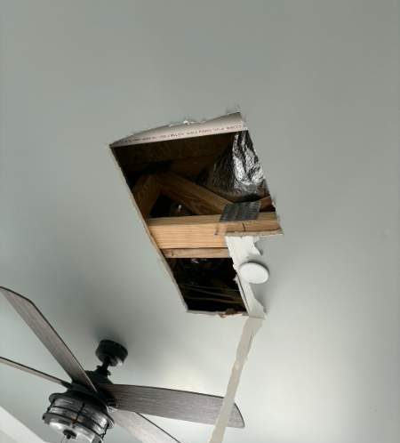 Rectangular ceiling opening with exposed framing beside a bedroom ceiling fan before repair